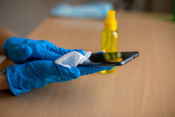 Woman's hand in blue gloves sanitizing cleaning smartphone mobile phone with alcohol on wood table surface with wet wipes