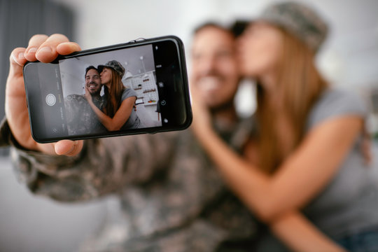 Young soldier taking selfie with his wife at home. Loving couple sitting on sofa in living room.