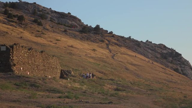 Old ruined building is located on mountain sunset or sunrise. Small silhouettes of people climbe up and down. Group of people stands at remains of an ancient fortress. Green trees grow on a rocky hill
