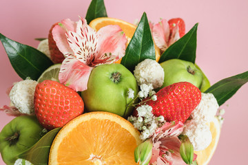 Close up of original unusual edible bouquet of fresh fruit isolated on pink background. Eating basket made of orange, kiwi, coconut, grape and candy. Decorated with green leaflets and coloring flower.