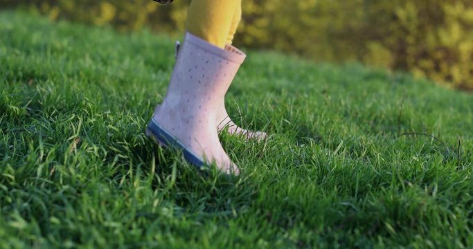 Children's legs stomp in pink rubber boots on green grass