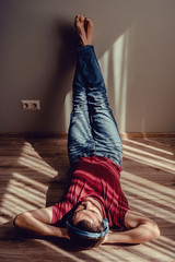 Carefree relaxed man wear jeans, wine red t-shirt listening to music lying down on parquet floor, feet on wall. Natural light and shadow pattern stripes. Stress free concept. Blue wireless headphones