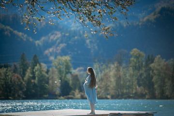 Pregnant girl with long hair in a blue dress on a bridge by the lake in the mountains in autumn or...
