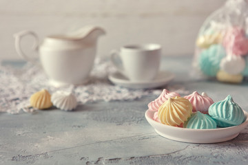Multi-colored meringues in pastel shades with cups and a jug on a light textured wooden background. The concept of Breakfast with tea or coffee, home comfort and tenderness. Close-up, selective focus