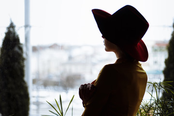girl in a dress and a hat looks out the window, sad, self-isolation, depression, pandemic, virus