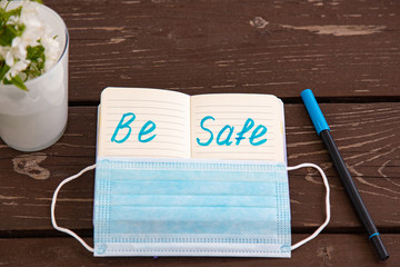 coronavirus be safe warning written in bright blue felt-tip pen in notepad with medical mask. brawn wooden table. view from above. flowers in a glass.