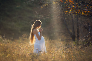 Pregnant girl with long hair in a white dress in the summer at sunset in the field prays