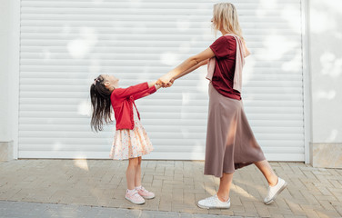 Side view image of beautiful mother looks to her little girl holds the hands together and dancing outside near their home. Daughter playing with her mom feeling happy. Woman and child shares love.