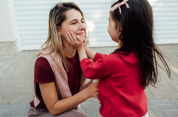 Closeup outdoor image of cute little girl embrace tender with hands her smiling mother on her face. Daughter hugs her mom after the preschool day. Happy woman hug her child next their house outside.