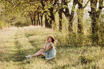 Naklejka na ściany i meble Young woman sitting on green grass and trees background, outdoors, wearing a vintage light green dress and a straw hat, on a sunny day.