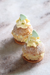 Contemporary key lime choux buns with craquelin, dipped in white chocolate, decorated with whipped diplomat cream and meringue cookies, on a marble board.