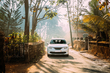 Goa, India. White Small Car Moving On Country Road Through An Indian Village. Morning Dawn Haze Enveloped Palm Trees