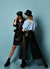 Fototapeta na wymiar Two young beautiful women in stylish black leather clothing, hats and massive boots standing over blue wall background