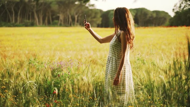 Girl in white striped dress photographs the wheat field with red poppies on the smartphone. Long hair woman walks around the beautiful countryside. Golden light in idyllic landscape. Spring. Summer.