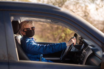 Woman in black face mask and sunglasses is driving a car and lookiing in the driver's opened window. Right-hand drive. Coronavirus infection control concept.