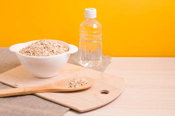 a white bowl with dry oatmeal, a bottle of water and a large wooden spoon on a cutting board, yellow background, copy space, top view, natural linen cloth under a bowl.
