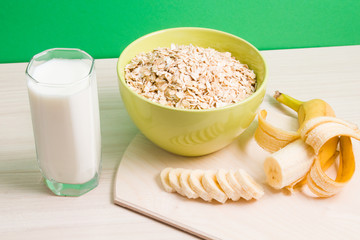 green bowl with dry oatmeal, staak of milk and sliced banana on wooden table, green background, proper nutrition concept, copy space.