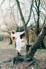 Naklejka na ściany i meble a young european blonde girl in a light white dress and black shoes stands on the root of a tree in a picturesque forest in autumn.natural,fashion concept