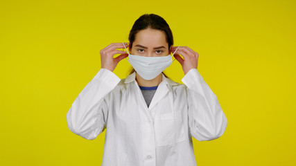 Young infectious disease doctor puts on a protective medical mask on her face. Girl in a white coat on yellow background. Coronavirus, flu