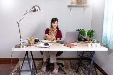 Pretty asian woman working remotely from home using laptop sitting on the couch or sofa in living room for work online with pet puppy cute dog and guardian, work life balance concept.