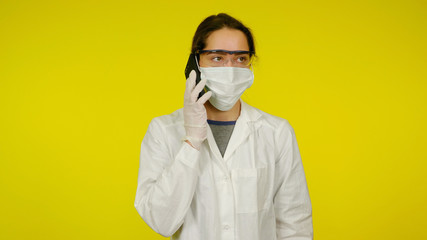 Young doctor in a medical mask, goggles and latex gloves is talking to patient on phone. Girl in a white coat on yellow background holds a smartphone near her ear. Coronavirus, flu
