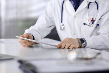 Unknown male doctor sitting and working with tablet computer in clinic at his working place, close-up. Young physician at work. Perfect medical service, medicine concept