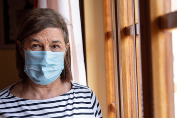 Ritratto di signora anziana che indossa una mascherina chirurgica  vicino alla finestra di casa sua