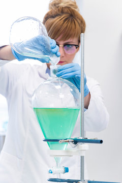 White female scientist wearing glasses and latex gloves works with green turquoise liquid in pear shaped separator funnel.