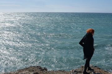 
A girl with red hair is standing with her back against the background of the sea. The sea sparkles in the sun