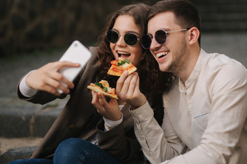 Young bloggers make selfie while eating slice of pizza. Beautiful couple in sunglasses smile to camera phone. Vegan fast food