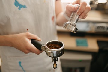 A female barista holds a metal tamper and a portafilter with coffee in a coffee shop. A barista preparing for brewing espresso or americano in a cafe.