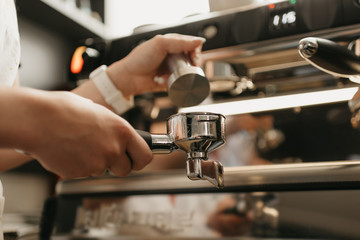 A close photo of female hands holding a metal tamper and a portafilter with coffee in a coffee shop. A barista preparing for pressing ground coffee for brewing espresso or americano in a cafe.