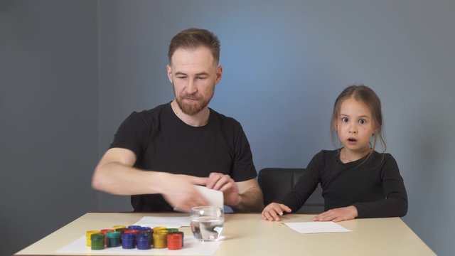 Dad teaches daughter to draw with color paints, Dressed in black on a gray background in the room