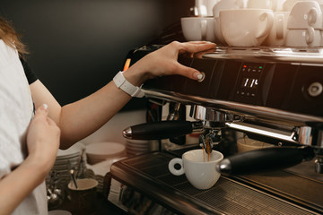 A female barista with white apron brewing espresso in a professional espresso machine in a coffee shop. A close-up photo of pouring a coffee in a white cup from a coffee machine in a cafe.
