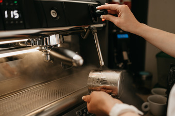 Fototapeta na wymiar A female barista wears a white apron purges a metal mug with steam in a professional espresso machine in a coffee shop. A close-up photo of cleaning a cup in a coffee machine in a cafe.