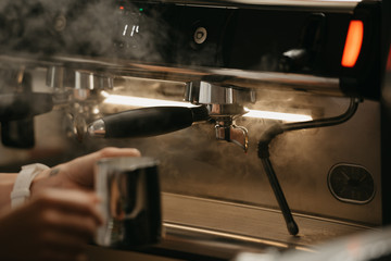 A female barista preparing to purge a metal mug with steam in a professional espresso machine in a coffee shop. A close-up photo of preparing to clean a cup in a coffee machine in a cafe.