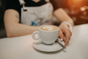 A female barista holds out a cup of fresh latte with a saucer and spoon to a client in a cafe. A barista preparing a customer order.