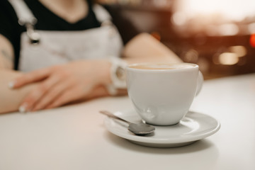 A female barista waits with a cup of fresh coffee with a saucer and spoon for a client in a cafe. A barista with a cup of latte in a coffee shop.