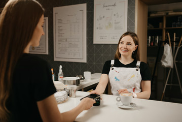 A female barista smiling holds out to a client a terminal for paying for a cup of coffee. A girl with long hair paying for a latte with a smartphone by contactless NFC technology in a cafe.