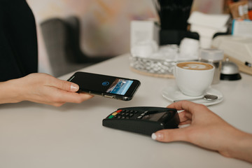 A girl paying for her latte with a smartphone by contactless NFC technology in a cafe. A female barista holds out a terminal for paying to a client in a coffee shop.
