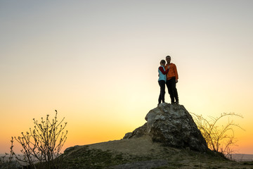 Man and woman hikers standing on a big stone at sunset in mountains. Couple together on a high rock in evening nature. Tourism, traveling and healthy lifestyle concept.