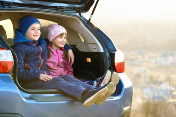 Two happy children boy and girl sitting together in a car trunk. Cheerful brother and sister hugging each other in family vehicle luggage compartment. Weekend travel and holidays concept.