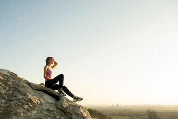 Fototapeta na wymiar Woman hiker sitting on a steep big rock enjoying warm summer day. Young female climber resting during sports activity in nature. Active recreation in nature concept.