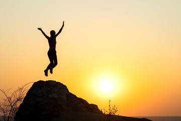 Silhouette of woman hiker jumping alone on empty rock at sunset in mountains. Female tourist raising her hands up standing on cliff in evening nature. Tourism, traveling and healthy lifestyle concept.