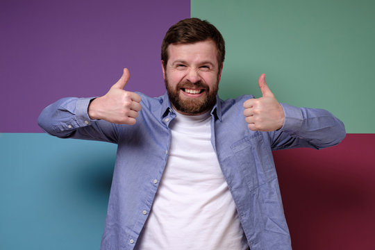 Gloating Caucasian man makes an insincere approving gesture with hands, raises thumbs up and grins teeth, squinting eyes.