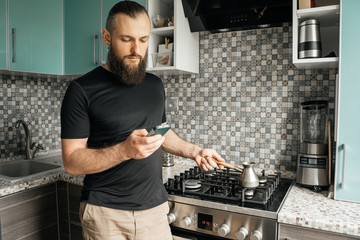 Attractive man with a beard in the morning looking at the phone brews coffee in cezve.