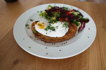 Poached egg on a piece of bread with green salad on a plate and on the wooden table. Healthy Breakfast