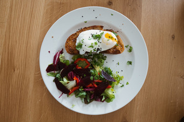 Poached egg on a piece of bread with green salad on a plate and on the wooden table. Healthy Breakfast