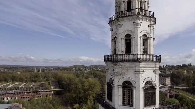 The old clock tower in the countryside area. Stock footage. Aerial view of the abandoned white tower surrounded by the city houses and green summer trees on blue cloudy sky background.