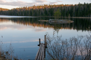 The old wooden pier on a lake in spring forest. Trees and sky reflected in the calm waters of a forest lake.  Natural background. Sunset moment. island in the lake. Stump. Sunset view. 